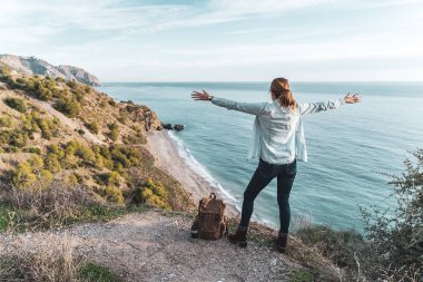 Young hip woman with open arms and a backpack explores the coast on a beautiful day. Concept of exploration and adventures