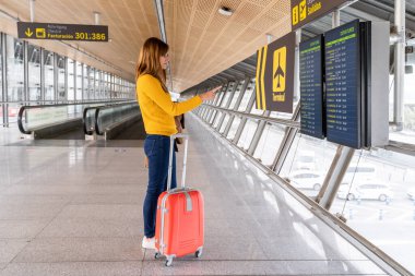 Beautiful young woman checking her flight's departure time on the information boards at the airport with her luggage. Travel and holiday concept.