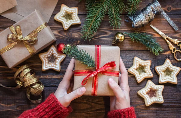 Dámské ruce drží dárek nad dřevěným stolem se smrkovými větvemi, perníčky a vánočními stromky. — Stock fotografie