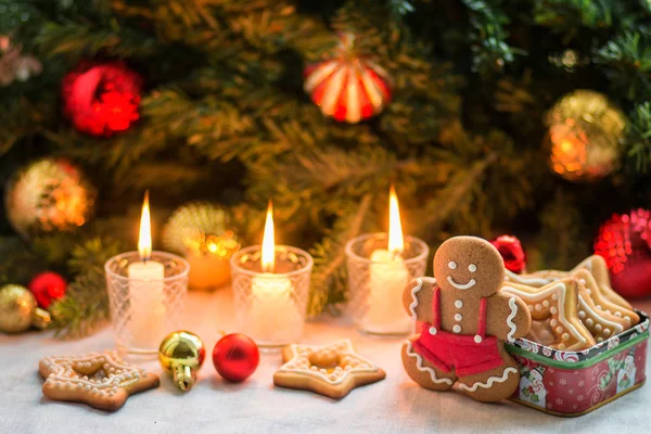 Noel kompozisyonu zencefilli adam, mumlar, küçük noel baloları, ev yapımı kurabiyeler ve beyaz yüzeyde bir hediye kutusu, arka planda Noel topları olan köknar dalları.  