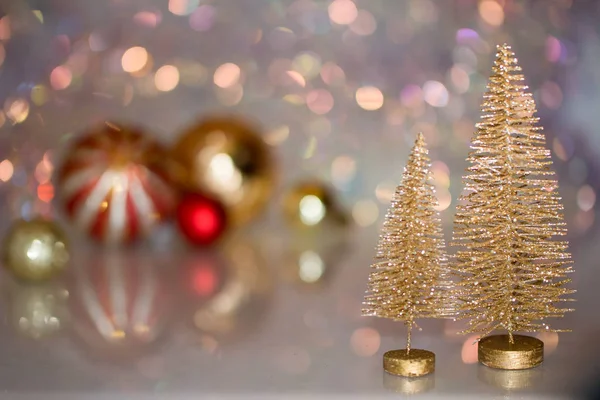Две маленькие золотые елки на стеклянной поверхности с разряженным фоном с боке и рождественскими шарами — стоковое фото