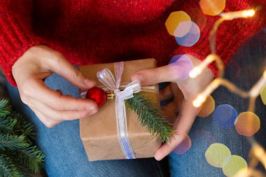 Kadın elleri hediye kutusunu beyaz kurdele, küçük kırmızı Noel topu ve köknar dallarıyla süslüyor. Çelenk ışıkları. Üst Manzara. Noel, yeni yıl, doğum günü konsepti 