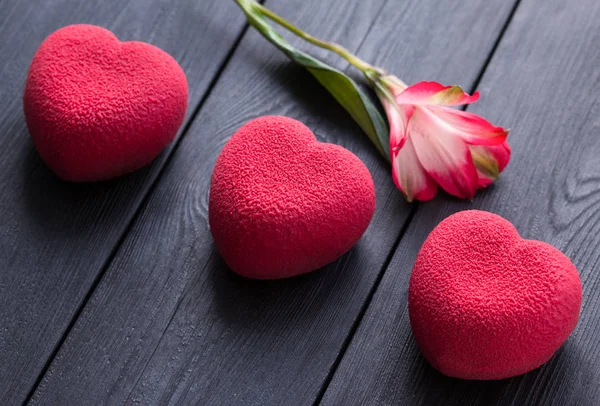 Concepto de San Valentín. Tres tortas de terciopelo corazón rojo sobre un fondo negro de madera con una delicada flor de Alstroemeria — Foto de Stock