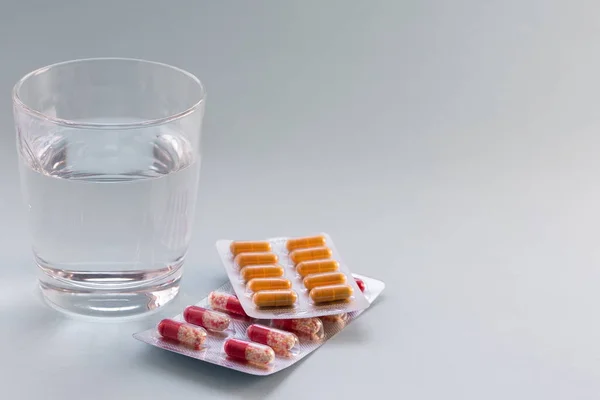 Cápsulas en blister con agua en una taza de vidrio transparente sobre una superficie gris. Concepto de medicina . — Foto de Stock