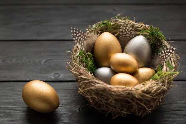 Gold and silver eggs with green moss and feathers in a nest on a black wooden table with copy space