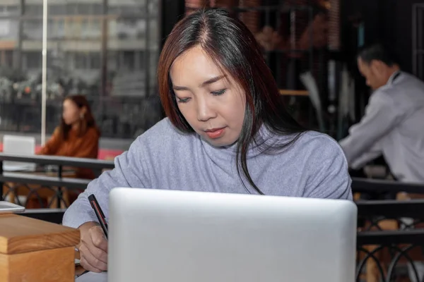 Smart beautiful clever Asian female write and note down information of summary positive study and business script essay in front of computer laptop at modern office table with the comfort atmosphere