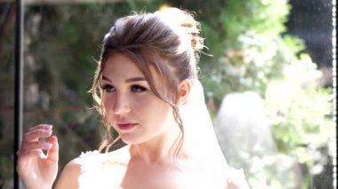 Saçları bukleli mutlu gelin camın yanında ağır çekimde duruyor.