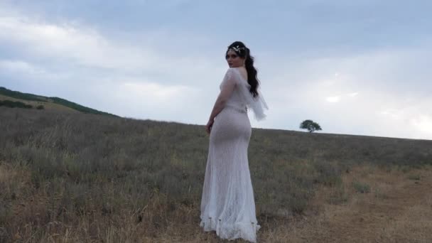 Dama en vestido blanco se para mirando hacia atrás contra el campo vacío — Vídeos de Stock