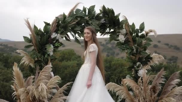 Elegant lady in long white wedding dress stands near arch — 비디오