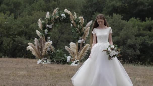花嫁はエレガントな結婚式のアーチに対して茶色の牧草地に沿って歩く — ストック動画