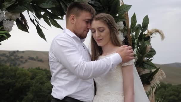 Verloofde knuffels bruid met vrolijke glimlach op bruiloft locatie boog — Stockvideo