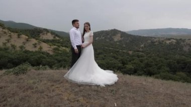bride holds handsome groom hand standing against hills