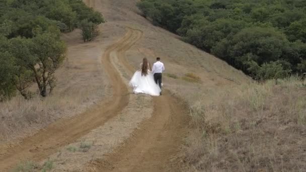 Para ślubna w eleganckim dressingu biegnie wzdłuż drogi gruntowej — Wideo stockowe