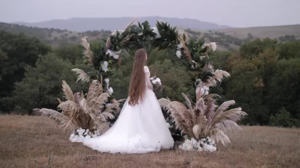 Υπέροχη νύφη με μακριά χαλαρά μαλλιά στέκεται στην αψίδα του γάμου — Αρχείο Βίντεο