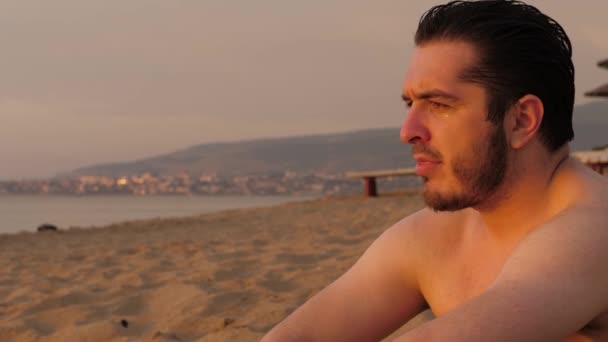 Bello uomo barbuto siede sulla spiaggia di sabbia gialla al tramonto — Video Stock