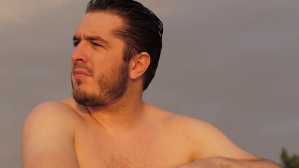 Ernster Typ mit schwarzen Haaren sitzt am Strand unter bewölktem Himmel — Stockvideo