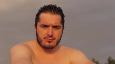 Çıplak gövdeli ciddi bir adam sahilde yakından bakar.