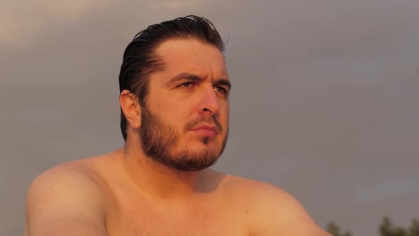 Guapo chico con torso desnudo y el pelo mojado se sienta en la playa — Vídeos de Stock
