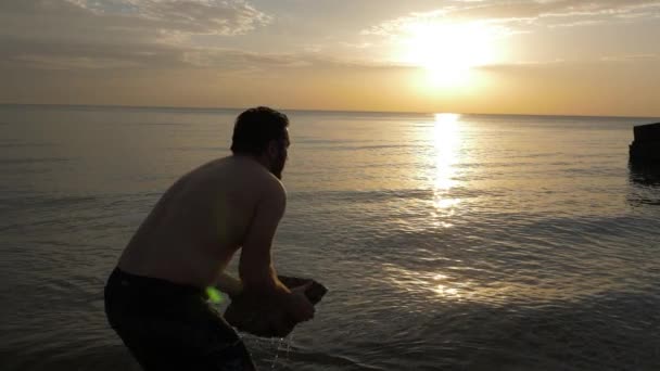 Silny mężczyzna z gołym tułowiem podnosi dużą skałę stojącą na morzu — Wideo stockowe