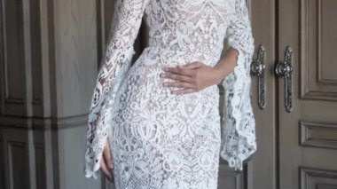 Beyaz dantelli gelinlik giymiş genç bir kadın kalçasında el gezdiriyor.