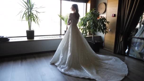 Невеста в длинном белом свадебном платье стоит у большого окна — стоковое видео