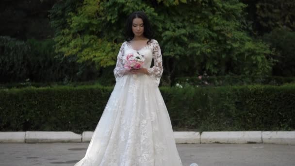 Hermosa chica en vestido de novia sostiene flores frescas en el parque — Vídeo de stock