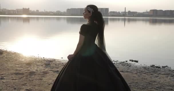 Брюнетка в длинном черном платье стоит на песчаном пляже — стоковое видео