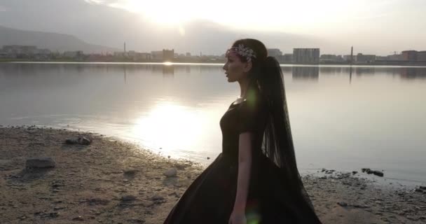 Dam i svart klänning med slöja står på sandstrand vid solnedgången — Stockvideo