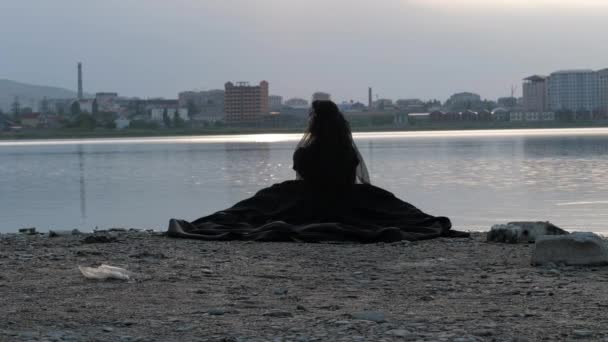 Леди в черном платье и прозрачной вуали сидит на берегу озера — стоковое видео