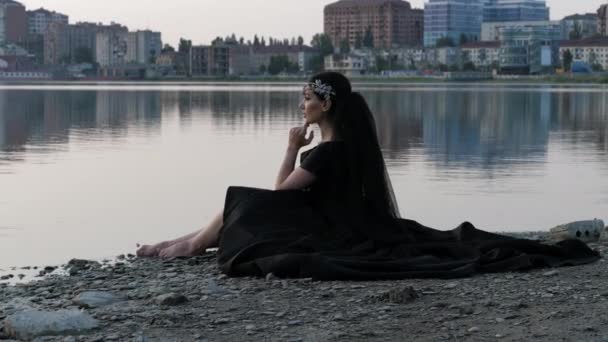 Босоногая всадница в длинном черном платье сидит на галечном пляже — стоковое видео