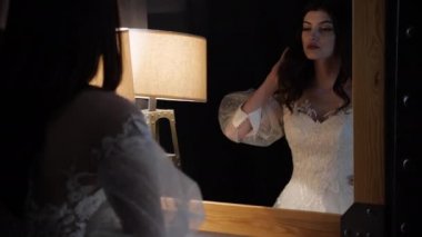 Beyaz, açık omuzlu esmer kadın aynaya bakıyor.