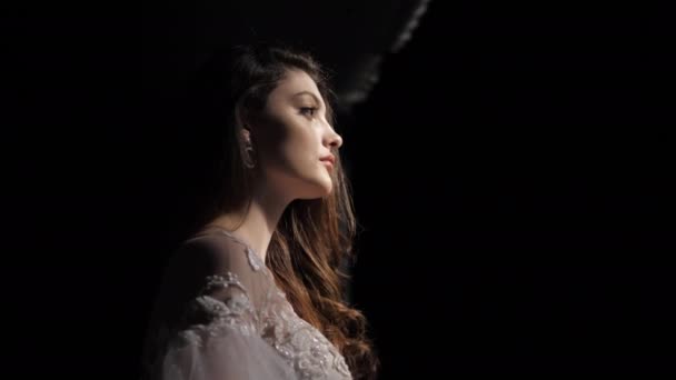 Элегантная брюнетка в белом стоит в темной комнате освещенной светом — стоковое видео