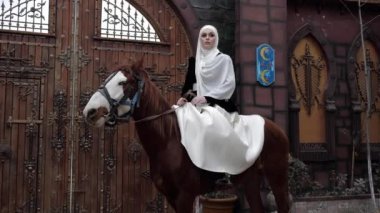 Başörtülü kadın kapının yanında oturmuş kestane atlarını okşuyor.