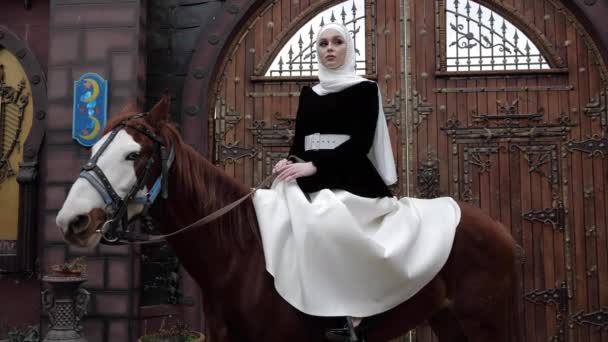 Arabic woman sits on chestnut horse near large wooden gate — Stock Video