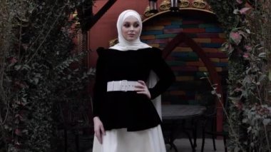 Shayla 'lı Arap elbiseli kadın yeşil çalıların yanında duruyor.