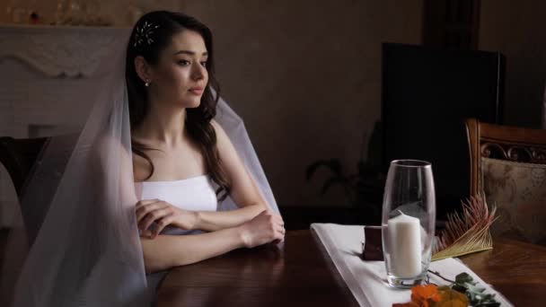 Σκεπτική νύφη με μακριά μαλλιά κάτω από το πέπλο κάθεται στο τραπέζι — Αρχείο Βίντεο