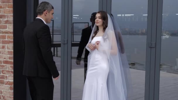 Mariée en robe longue se tient près de grandes fenêtres et fiancée câlins — Video