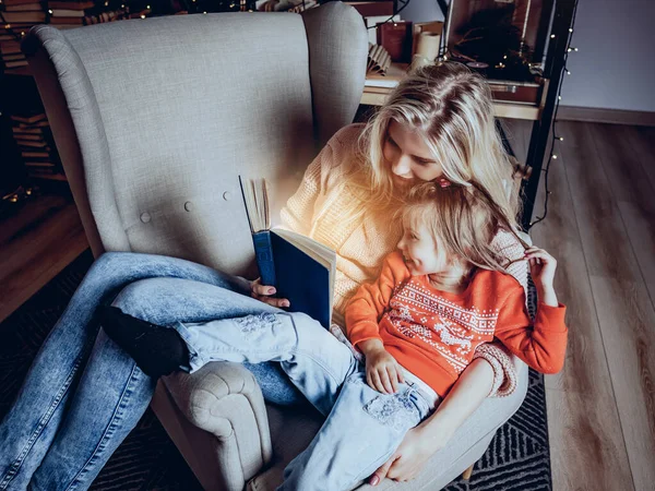 mom reads a book to her daughter sitting in a chair