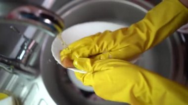 Sarı koruyucu eldivenlerle kadın ellerini kapat. Mutfak lavabosu köpük ve sofra takımlarıyla dolu beyaz tabağı mavi temizleme süngeriyle yıka.