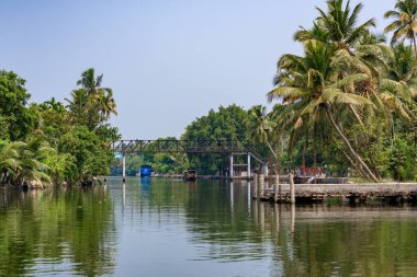 Alleppey, Kerala, Hindistan - 30 Mart 2018: Palmiye ağaçları ve demir köprüsü olan durgun kanal. Sabah küçük kırmızı bir tekne ters yöne giderken çekilmiş.