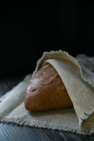 Siyah arka planda keten peçeteye sarılı organik buğday ve çavdar unundan yapılmış taze siyah ekmek. Sağlıklı bir kırsal kahvaltı için taze hamur işleri..