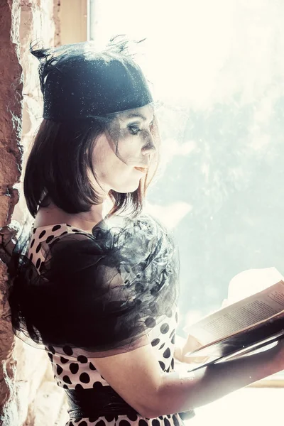 Geçen yirmili yılların tarzında giyinmiş siyah şapkalı, duvağı açık omuzları açık, elinde kitapla pencerenin kenarında duran bir kız..