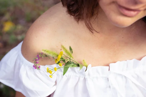 Attractive woman in a white blouse with an open shoulder and a bouquet of wildflowers in the neckline. A slight smile on the lips of a young beauty with dark hair.