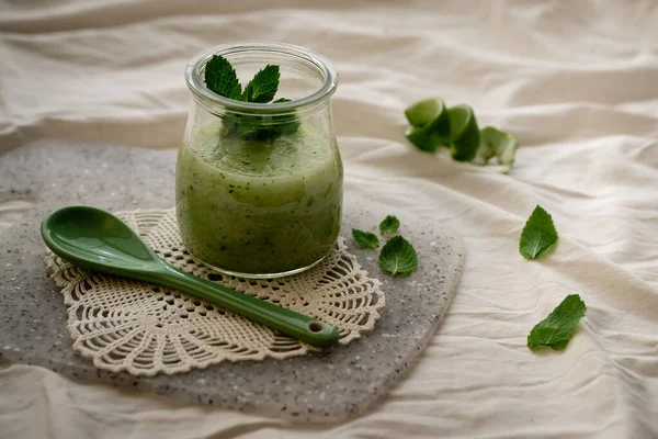 Yeşil porselen kaşıkla cam bir kavanozda meyve püresi. Nane yaprakları ve limon kabuğuyla açılan bir ağda.