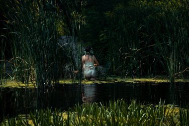 30-40 yaş arası, açık renkli bir yaz elbisesi giyen genç bir kadın kumlu sahilde sazlıklarla birlikte yosunlarla sudan çıkar.