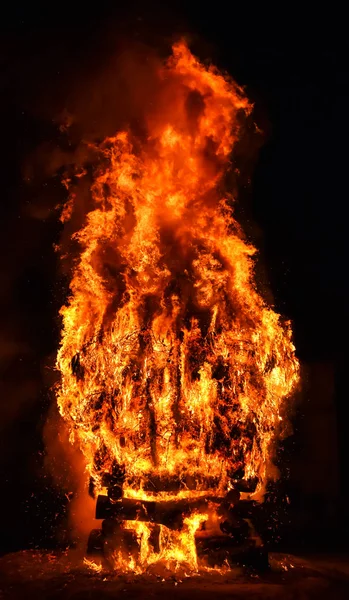 Incredibile violento falò di fuoco in un villaggio nella notte buia. La combustione della legna da ardere crea grandi fiamme, razzi, fumo, gas e genera un'ampia luce intorno alla piazza. Foto verticale . — Foto Stock
