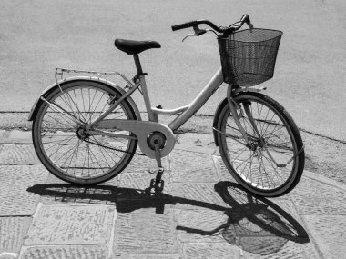 Bir şehir sokağının kaldırımına gölgesini atan bir bisikletin siyah beyaz fotoğrafı. Olay yerindeki hiç kimse aracı kullanmıyor. Bisikletin gidonunda siyah bir sepet var..