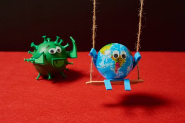 Conceptual image of a toy green virus behind a toy earth that is going to catch to the earth that is unprepared having fun on a swing on a black and red background. clipart