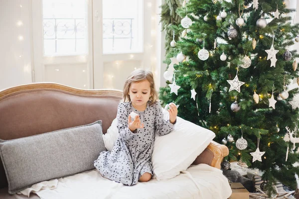 La fille est assise sur le canapé et mange des bonbons à côté du sapin de Noël décoré, la chambre est décorée pour la nouvelle année et Noël — Photo