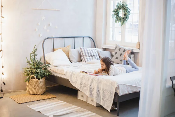 La chica está acostada en el sofá y leyendo un libro, la habitación está decorada para el nuevo año y la Navidad —  Fotos de Stock